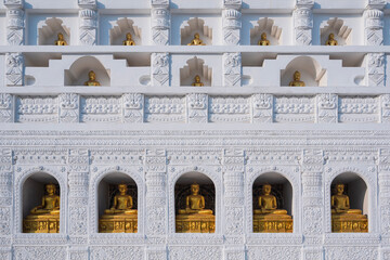 Fototapeta premium Golden Buddha statues in white niches of a Thai temple wall. Traditional Buddhist architecture with intricate carvings and repeating patterns. Spiritual and cultural landmark in Thailand.
