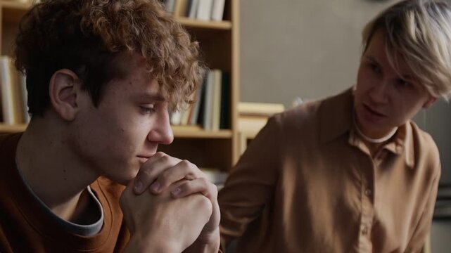 Distressed teenage boy describing feelings to female therapist during group discussion then hiding face with hands struggling to cope in counselling room