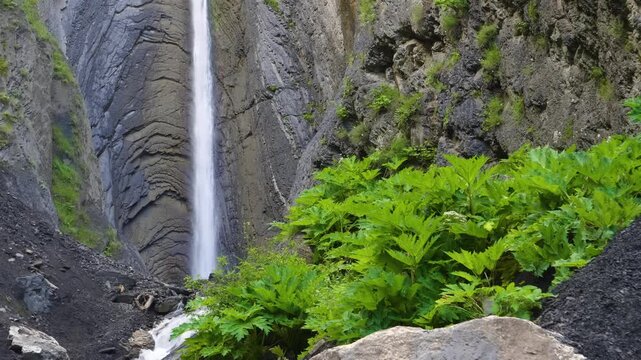 A scenic view of the powerful Zruh Waterfall as it cascades gracefully over stratified cliffs, surrounded by lush green foliage and rugged rocks. The footage captures the natural beauty and tranquilit