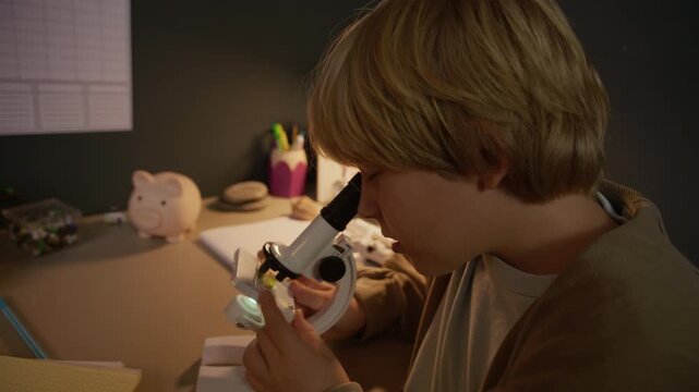Close up slowmo of inspired Caucasian scientist boy sitting at well-organized desk and observing leaf specimen under microscope preparing for STEM class at home