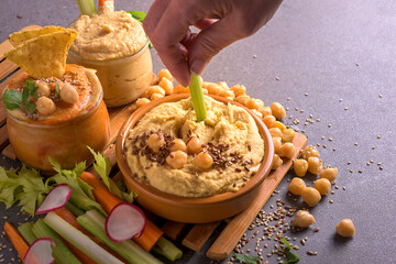 Hand of a woman dipping a fresh vegetable stick into creamy hummus bowl with seeds and chickpeas