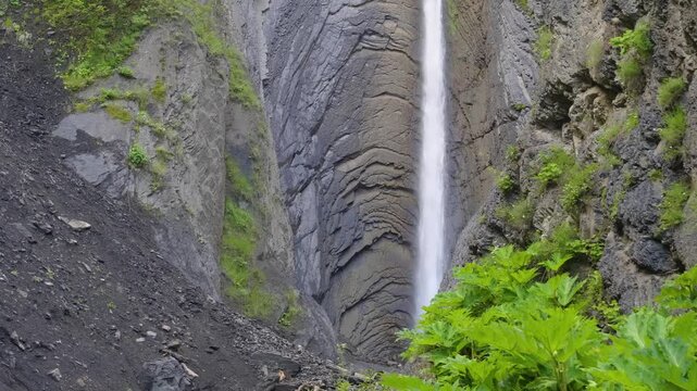 The beautiful, tall Zrykhskiy Vodopad waterfall cascades through a dramatic V-shaped gorge amidst the rugged green mountain landscape of Dagestan, Russia, in the summer. The footage captures the raw p