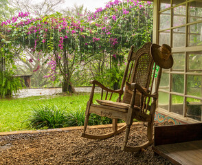 A vintage wooden rocking chair with a straw hat rests on a porch in a lush garden setting in Costa Rica. The scene evokes relaxation and vacation