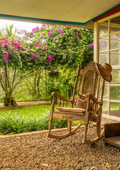A vintage wooden rocking chair with a straw hat rests on a porch in a lush garden setting in Costa Rica. The scene has relaxation and vacation themes