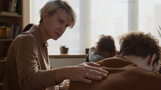 Middle aged female psychotherapist giving supporting touch to young boys shoulder during emotional outburst in group session for adolescents