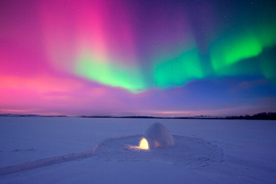 A lonely igloo glowing from inside in a frozen snow field with Aurora borealis in night sky. Northern lights in winter field. Wintry scene with glowing polar lights and snowy igloo. Christmas postcard - Powered by Adobe