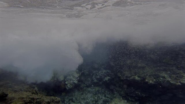 Underwater waves hitting a brown rock formation causing white foam and bubbles in blue ocean 