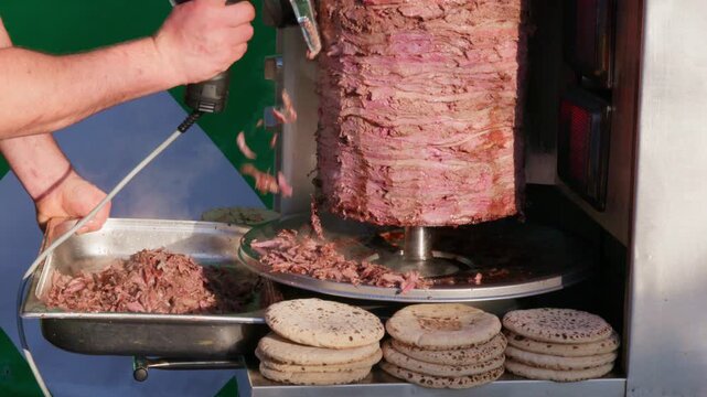 Street vendor slices hot shawarma doner from a vertical rotisserie with an electric knife as meat falls into a tray, fresh pita breads stacked in front, ready for fast service