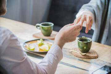 Man putting engagement ring on smiling woman's finger during romantic proposal at home with coffee