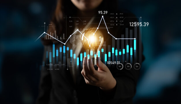 A businesswoman holds a light bulb surrounded by digital financial data graphics, symbolizing innovation, strategy, and analytical thinking in a modern workspace. Scalp - Powered by Adobe