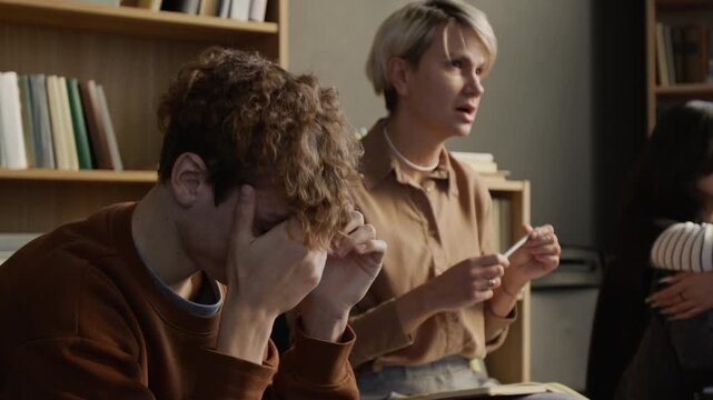 Distressed teenage boy covering face with overwhelmed expression during group discussion then female therapist attentively assessing students emotional state in counselling room