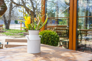 Bouquet of fresh spring flowers including yellow tulips arranged in white metal vase on wooden table in outdoor cafe with blooming trees. Warm light, garden reflections, and seasonal floral decoration