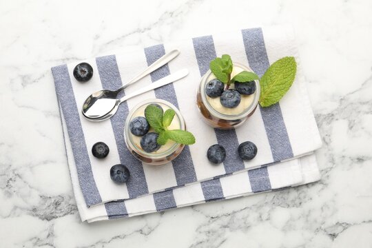 Tasty trifle dessert with whipped cream, mint and blueberries in glass jars on white marble table, flat lay