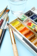 Palette with watercolor paints, tubes, glass of water and brushes on light blue background, closeup