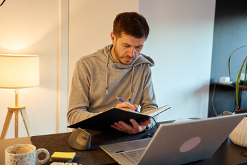 Man standing at home office desk writing in notebook with pen next to laptop and desk lamp