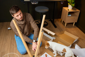Man assembling wooden lamp structure while dog stands next to him with unpacked boxes