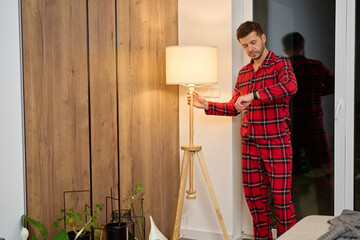 Man in red pajamas standing by floor lamp and checking wristwatch in living room