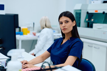 Medical professional in a lab setting working on a computer while a colleague conducts tests in the background