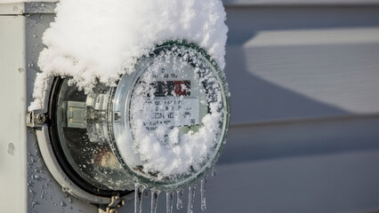 Thick snow on electric meter, covering face and top, with icicles hanging below. Accumulated snow on electric meter shows winter weather impact on utilities.