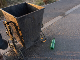 Metal trash can on a sidewalk with a plastic bottle lying on the ground. Littering and...