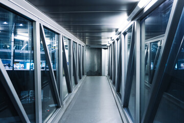 Empty glass jet bridge corridor at airport terminal. Modern passenger boarding walkway interior. Symmetric perspective tunnel with lights. Travel infrastructure at night. No people inside.