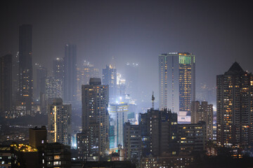Naklejka premium Dense urban skyline at night with illuminated high-rise buildings emerging through atmospheric haze, representing modern metropolitan city life in India