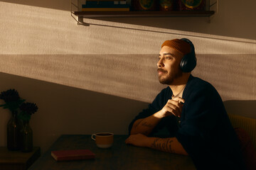 A peaceful man wearing headphones sits at a kitchen table, enjoying music or a podcast in the warm glow of sunset. The golden hour light creates a serene atmosphere in a cozy home environment. © Francis