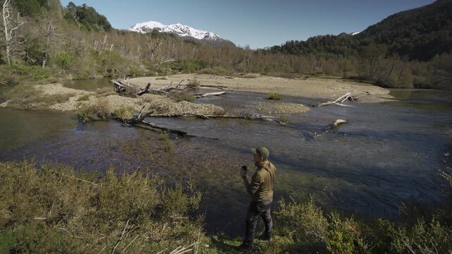 Caucasian man drinking water from the Pichi Traful River using a cup with snow-capped mountains and lush vegetation in the background, Patagonia