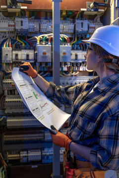 Woman electrician compares paper schematics with switchgear. Manual audit.
