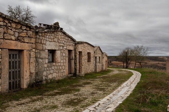 Traditional stone wine cellars in Moradillo de Roa, Burgos, Spain, lining a curved rural path under an overcast sky. A rustic heritage landscape showcasing historic architecture and countryside scener