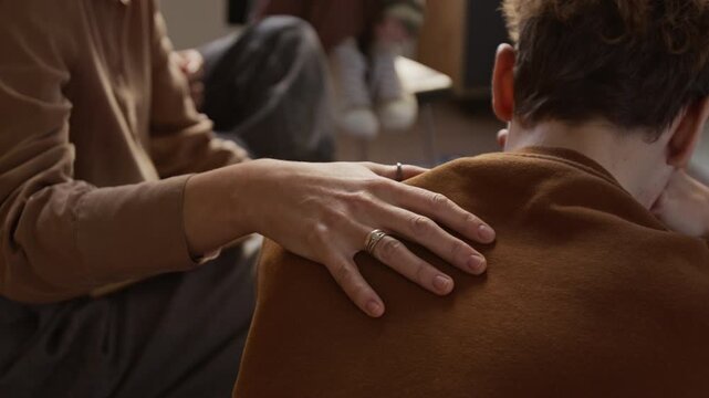 Close up on hand of female psychotherapist resting gently on teenage boys shoulder offering reassurance during emotional moment in therapeutic group session for adolescents