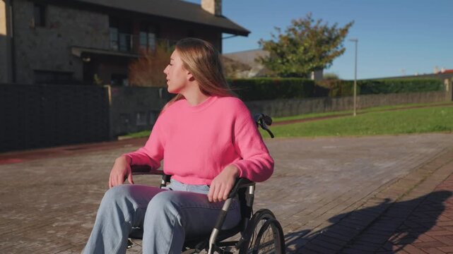 Young disabled woman sitting in her wheelchair outdoors in a residential area, enjoying the sun and looking around her as she moves along the sidewalk on a beautiful, bright day