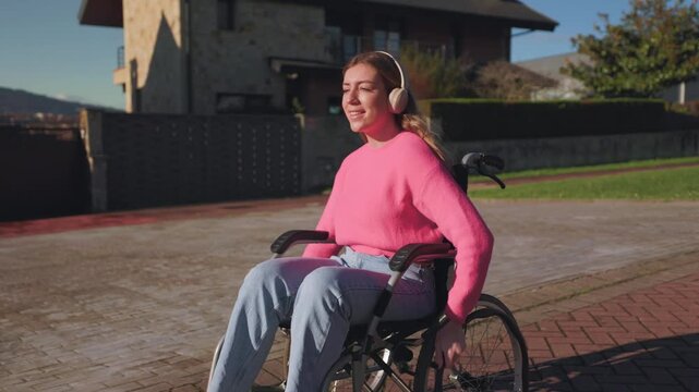 Beautiful young paraplegic woman wearing headphones while propelling her manual wheelchair along a sunny residential street, enjoying a moment of freedom, independence, and happiness