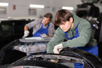 Focused young auto body technician in casual olive hoodie and blue overalls carefully repairing car...