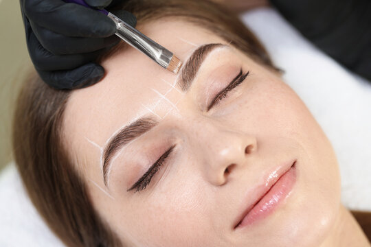 Beautician drawing marks on woman's face before permanent eyebrow makeup procedure indoors, closeup