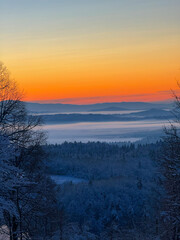 Naklejka premium Soft morning light glows above a foggy valley as snowy forests stretch toward distant hills. Contrast between warm sunrise colours and cool winter tones creates a serene and timeless winter landscape.