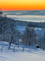 Naklejka premium Snowy hills descend into a quiet valley wrapped in soft mist as the sun rises. Warm colours on the horizon contrast with cool winter blues. Peaceful rural landscape reminiscent of a winter fairy tale.