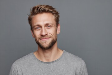 Portrait of a relaxed fair haired man in a t shirt smiling at the camera with confidence against a gray backdrop with space on the right