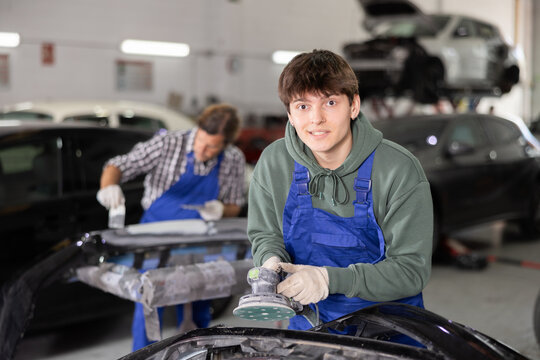 Positive young man in comfortable green hoodie and blue overalls working in auto body repair shop, expertly sanding car parts with sander, preparing for painting