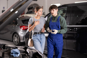Professional young auto technician explaining required car engine maintenance to interested female customer while standing next to open hood at vehicle repair shop ..