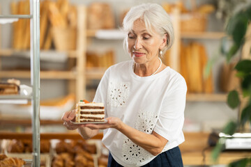 Pleased old female customer holding pastry on paper plate in bakery shop