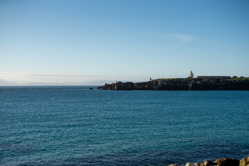 Calm blue sea with rocky coastline and distant lighthouse under clear sky creating peaceful coastal landscape with open water horizon and natural maritime atmosphere