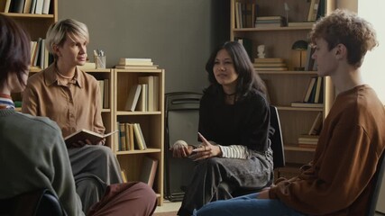 Teenage girl holding baseball speaking to group of fellows in guided therapy session with female therapist while others actively listening, participants using object to take turns in counselling room
