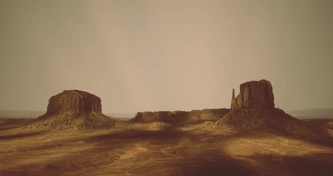 Sunlight bathes the serene desert landscape as two impressive rock formations tower above the warm, earthy terrain. Shadows stretch across the ground, enhancing the natural beauty of the scene.