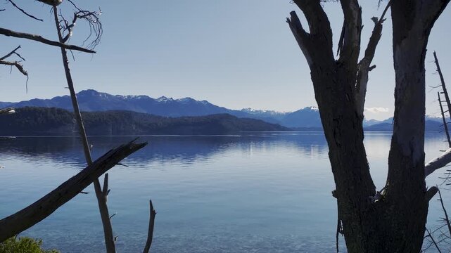 Man with a backpack standing by a calm Nahuel Huapi Lake looking at a mountain range through dead trees in Patagonia