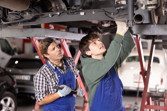 Two focused professional mechanics, middle aged and young men, wearing blue overalls, repairing undercarriage of car raised on lift in auto repair shop..