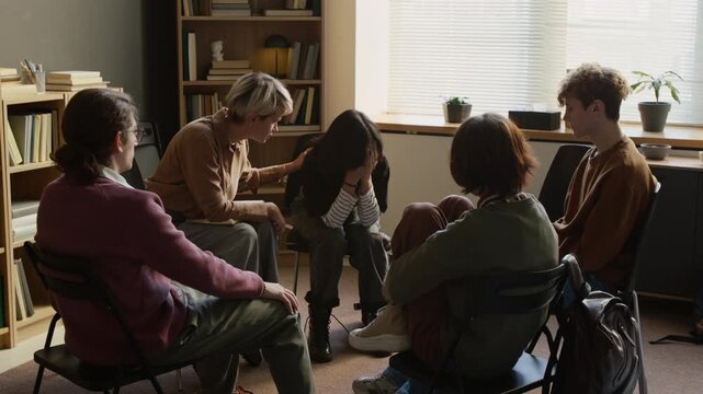 Female psychotherapist comforting crying teenage girl having emotional breakdown during group therapy then resistant adolescent leaving counselling room