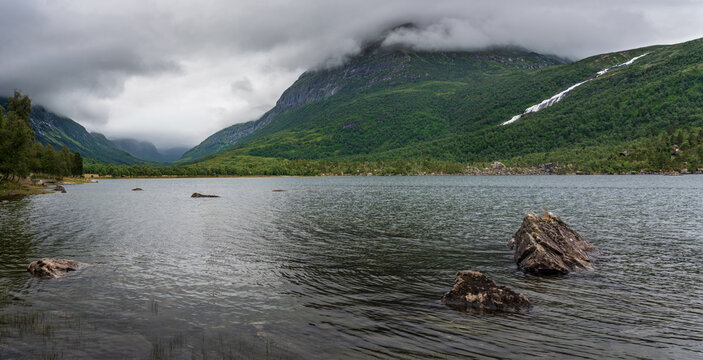 The scene shows Innerdalen nature reserve in Norway, featuring Innerdalsvatna lake, the cloud-covered Innerdalst&aring;rnet mountain peak, and a waterfall in the distance.
