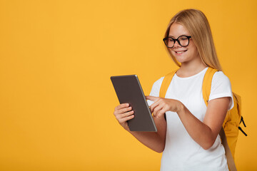 Smiling adolescence caucasian schoolgirl in white t-shirt, glasses with backpack studies with tablet, isolated on yellow background. Modern education with gadget, remote lesson class, social distance