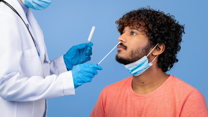 A healthcare worker in a white coat performs a nasal swab test on a male patient. The patient is...